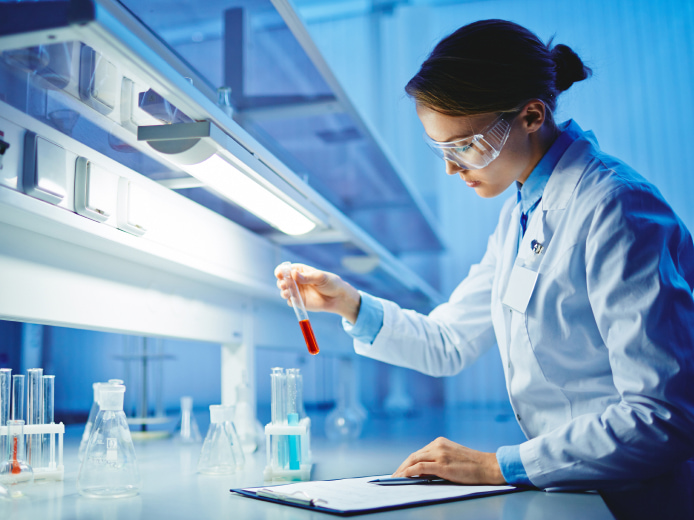 Focused scientist examining a test tube with red liquid in a modern lab setting, with notepad and lab equipment in view.